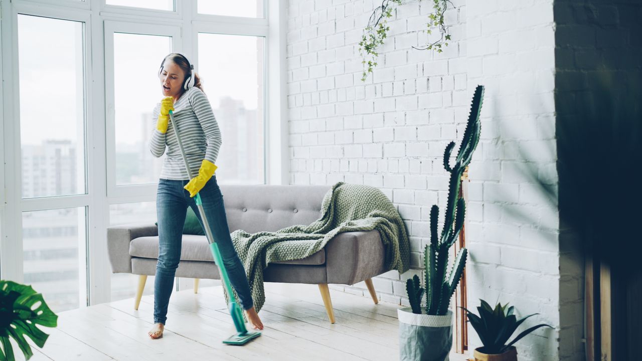 Cheerful woman with headphones singing and cleaning the living room with a mop.